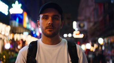 Erkek gezgin doğrudan izleyiciye bakıyor, neon ışıkların altında hareketli bir caddede duruyor.
