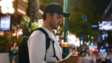 Sırt çantalı bir adam akıllı telefon kullanıyor, geceleri şehir sokaklarında geziniyor.