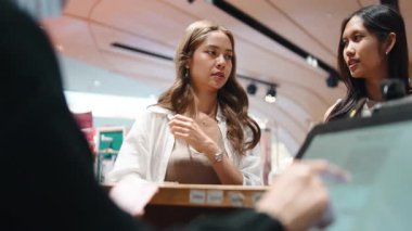 İki bayan arkadaş alışveriş merkezinin kasasında bir satıcıyla konuşup etkileşime geçiyorlar.