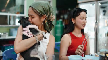 Genç Asyalı bir kadın arkadaşlarıyla bir restoranda sevgi dolu bir köpeğe sarılıp sarılıyor.