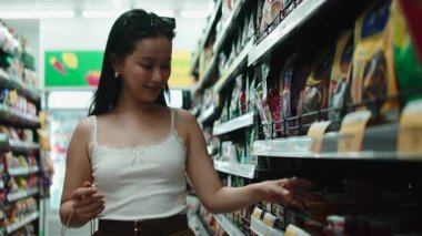 Yiyecek almak için alışverişe çıkan genç bir kadın, bir mağaza koridorundaki raftan bir ürün seçiyor.