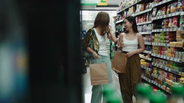 İki bayan arkadaş birlikte yürüyor ve bir markette raftan ürünleri seçiyorlar.
