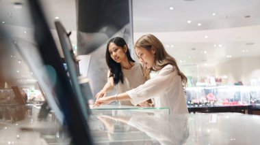 Perakende terapiden zevk alan iki Asyalı kadın cam vitrinlerden eşyalar seçiyor.