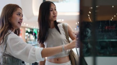 İki Asyalı bayan arkadaş bir mağaza vitriniyle etkileşime geçip, sıradan bir alışveriş deneyiminin keyfini çıkarıyorlar.