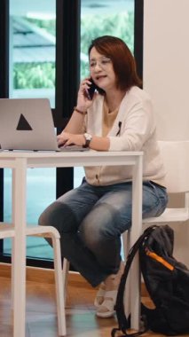 40 'lı yaşlarda Asyalı bir iş kadını modern bir ofiste telefonla konuşurken dizüstü bilgisayarla çalışıyor.