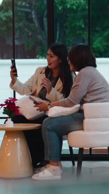 Asyalı anne ve kızı rahat bir oturma odasında otururken akıllı bir telefonla mutlu bir şekilde selfie çekiyorlar.