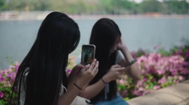 İki genç Asyalı kadın gölün kenarındaki taş bir bankta oturmuş akıllı telefonlarıyla fotoğraf çekiyor ve gülüyorlar.