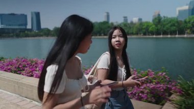 İki genç kadın göl kenarında yürürler. İkisi de çiçeklerle ve modern binalarla çevrili akıllı telefonlarının ekranına bakarlar.