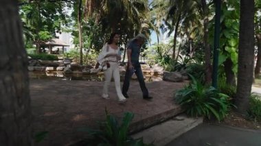 Güneş gözlüğü takan Asyalı bir adam göl ve ağaçların yanındaki kayalık kaldırımda elinde bir şişe su tutan beyazlı bir kadının yanında yürüyor.