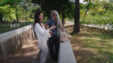 Uzun beyaz saçlı Asyalı bir adam parktaki köprü duvarına tripod yerleştiren bir kadının yanına.