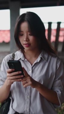 Asyalı kadın dışarıda akıllı telefonuyla fotoğraf çekerken gülümsüyor.