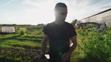 Uzun saçlı, bin yıllık beyaz çiftçi elinde tabletle gün batımında şehir bahçesinde geziniyor - 4K Yatay