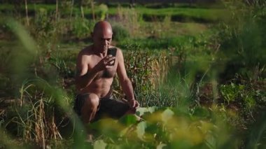 Zengin bir şehir bahçesinde çömelmiş kel bir adam sebzelerinin fotoğrafını çekmek için akıllı telefonunu kullanıyor - 4K Yatay