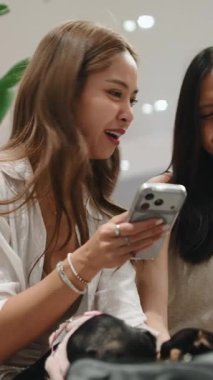 Köpekleri alışveriş merkezinde bir çantada dinlenirken iki Asyalı genç kadın akıllı telefon kullanıyor.