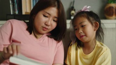 Genç Asyalı anne ve kız, modern evlerinde rahat bir kanepede kitap okuyarak birlikte iyi vakit geçiriyorlar.