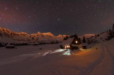 Yüksek Tatralar 'da gece kışı manzaraları, arka planda karla kaplı dağlar ve yıldızlı gökyüzü olan dağ evleri.