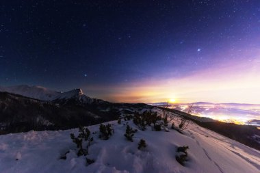 Yüksek Tatralar 'da gece kışı manzaraları, arka planda karla kaplı dağlar ve yıldızlı gökyüzü olan dağ evleri.