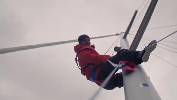 Активный мужчина с улыбкой лазает по мачте на белой спортивной яхте с российским флагом под низким углом — стоковое видео