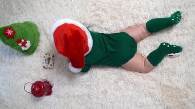 Noel Baba şapkası takan ve yeşil Noel elbisesi giyen, süslü püslü bir battaniyeye uzanmış, küçük bir fener, paketlenmiş bir hediye ve bayram süsü. Bebek elf yuvarlanıyor