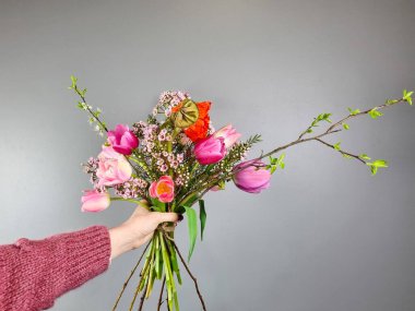 Pembe laleli bir buket, çiçek açan dallar ve tarafsız gri arka planda kır çiçekleriyle el ele tutuşmak. Çiçekçi dükkanında çalışıyor.