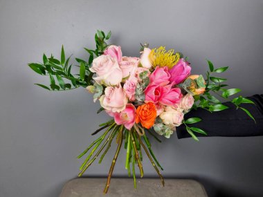 Renkli çiçek buketi pembe güller ve yemyeşil çiçeklerle düzenlenmiş, tarafsız gri arka planda sergileniyor. Çiçekçide çiçekçi