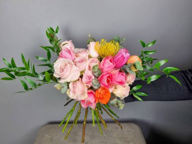 Renkli çiçek buketi pembe güller ve yemyeşil çiçeklerle düzenlenmiş, tarafsız gri arka planda sergileniyor. Çiçekçide çiçekçi