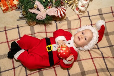 Noel Baba kostümü giymiş bir bebek Noel ağacının altında rahat bir battaniyenin üzerinde yatıyor ve elinde Noel süsü olarak bir balo süsü tutuyor. Noel ağacını süsle