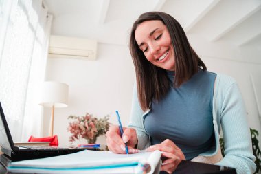 Dizüstü bilgisayarla donatılmış bir masada otururken not defterinde kalemle notlar yazan odaklanmış genç bir kadın. Gülüşü öğrenmenin verdiği hazzı ve bir öğrencinin adanmışlığını yansıtıyor.