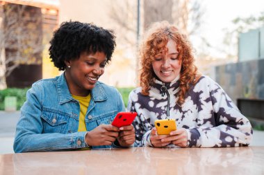 İki neşeli genç kadın hevesle akıllı telefonlarını kullanıyor, mobil uygulamalarla uğraşırken keyifli bir deneyim paylaşıyorlar, teknolojiye ve birbirleriyle bağlantıya dalmışlar..