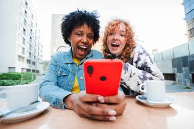 İki neşeli genç kadın, kafeterya masasında birlikte akıllı bir telefona baktıklarında, bu canlı ortamda gerçek mutluluk ve bağlantı ifadelerini sergilerken heyecanlı bir şekilde keyifli anlar paylaşıyorlar.