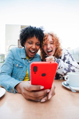 İki neşeli genç kadın, bir akıllı telefonla iletişim kurarken heyecan ve mutluluğu ifade ediyor, bir mobil uygulama ile etkileşim kurarken kahkaha ve bağlantılarla dolu keyifli bir anı paylaşıyorlar..