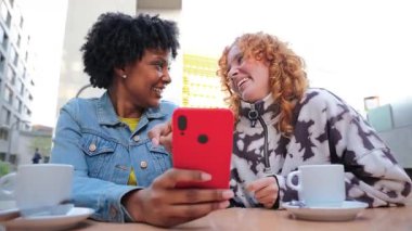 İki neşeli genç kadın, kafeterya masasında birlikte akıllı bir telefona baktıklarında, bu canlı ortamda gerçek mutluluk ve bağlantı ifadelerini sergilerken heyecanlı bir şekilde keyifli anlar paylaşıyorlar.