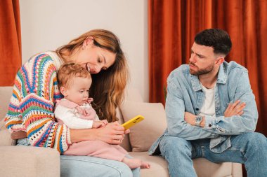 Bir anne, küçük kızı yanında oynarken akıllı telefonuna odaklanır, babasının somurtmasıyla gözlemlediği gibi, dijital çağdaki aile etkileşimlerinin dinamiklerini vurgular..