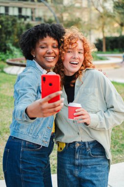 İki hevesli kız, mutlu bir şekilde cep telefonuyla keyifli bir selfie çekerken açık havada neşeli bir şekilde kahkaha ve neşe paylaşıyorlar, arkadaşlığı ve iletişimi kutluyorlar..