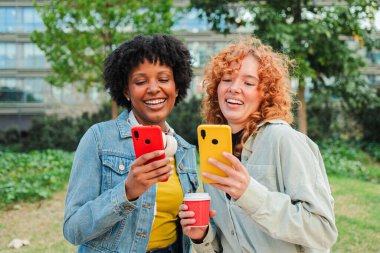 İki neşeli genç kadın birlikte gülerken ve anlarını paylaşırken akıllı telefonlarıyla meşgul oluyorlar, parlak renkli telefon kılıflarını sergiliyorlar ve mobil uygulamaları kullanma tecrübelerini paylaşıyorlar..