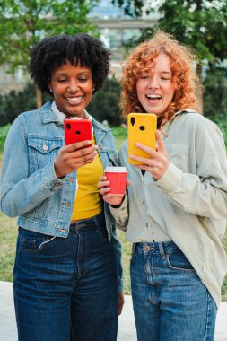 İki neşeli genç kadın akıllı telefonlarıyla meşgul, güneşli bir açık alanda kahvelerini yudumlarken keyifli bir anı paylaşıyorlar, teknoloji ile dostluk ve bağlantı gösteriyorlar..