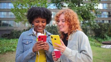 İki neşeli genç kadın birlikte gülerken ve anlarını paylaşırken akıllı telefonlarıyla meşgul oluyorlar, parlak renkli telefon kılıflarını sergiliyorlar ve mobil uygulamaları kullanma tecrübelerini paylaşıyorlar..