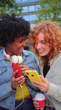 İki neşeli genç kadın akıllı telefonlarıyla meşgul, güneşli bir açık alanda kahvelerini yudumlarken keyifli bir anı paylaşıyorlar, teknoloji ile dostluk ve bağlantı gösteriyorlar..