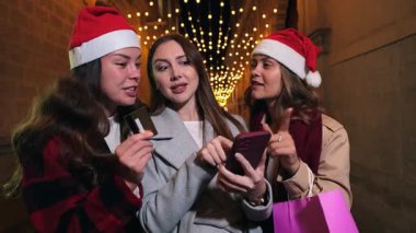 Noel Baba şapkalı genç kadınlar akıllı telefon ve kredi kartıyla online alışveriş yapıyorlar, tatil satışları, dijital ödemeler, dekoratif ışıklar altında bir akşam gezisinde Noel deneyimleri sergiliyorlar.