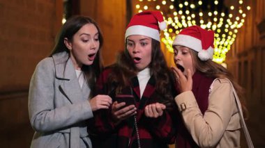 Akıllı telefonlarıyla hediye arayan neşeli bir arkadaş grubu, Noel kutlamaları sırasında heyecanı paylaşıyor, arkadaşlığın ve bayram neşesinin ruhunu yakalıyorlar..