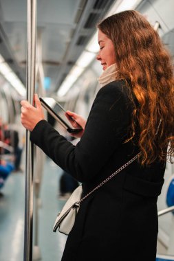 Odaklanmış genç bir kadın metroda seyahat ederken tablet okuyarak eğleniyor. Bu an, yolcuların yoğun seyahat deneyimi sırasında teknolojinin ve eğlencenin karışımını yakalar.