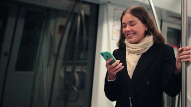 Cep telefonuyla meşgul neşeli bir bayan, metro yolculuğu sırasında sosyal medya etkileşimlerine dalmış, modern teknolojinin günlük yaşamdaki bağlantısını ve bağlılığını gözler önüne seriyor.