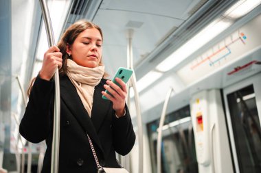 Metroda seyahat ederken akıllı telefonunu kullanan, seyahati sırasında teknolojiye olan bağlılığını vurgulayan, mobil uygulamaların günlük seyahat entegrasyonunu gösteren odaklanmış bir kadın.