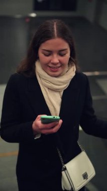 Neşeli genç bir kadın akıllı telefonuyla meşgul olurken hareketli bir ulaşım merkezinde yürüyen merdiven çıkıyor ve günlük hayatta teknoloji ve bağlantıyla neşeli etkileşimini gösteriyor..