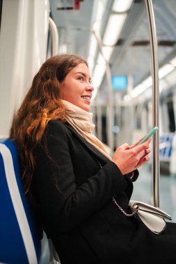 Metroda otururken akıllı telefonunu kullanmakla meşgul neşeli genç bir kadın, cep telefonundaki farklı uygulamalara dalmış, keyif ve bağlantıyı yansıtıyor.