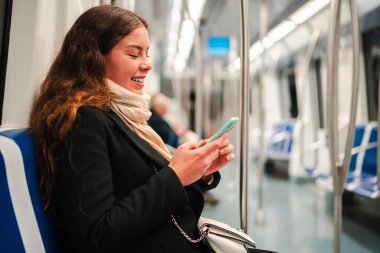 Mutlu bir bayan yolcu akıllı telefonuna daldı, çeşitli uygulamaları keşfetti ve metroya binerken internete göz attı, günlük seyahat rutininde teknolojinin keyfini gösterdi.