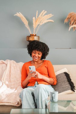 Evdeki koltukta rahat bir şekilde otururken akıllı telefonunu kullanmakla meşgul neşeli bir kadın. Kişisel alanından memnun ve gülümsüyor. Bir anlık rahatlık ve bağlantı sergiliyor..