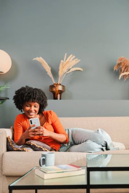 Neşeli kız rahat bir oturma odasında koltukta dinlenirken cep telefonuyla meşgul oluyor, evde vakit geçiriyor ve akıllı telefonuyla uygulamaları zevkle keşfediyor..