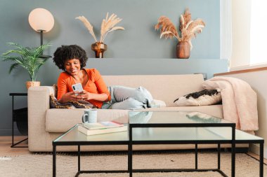 Keyifli bir kadın rahat bir kanepede uzanıyor, akıllı telefonuyla derinden meşgul, evine davet ederek rahatlama ve eğlencenin tadını çıkarıyor. Konfor ve teknolojinin mükemmel bir karışımı..