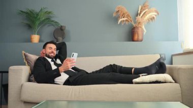 Genç bir iş adamı rahat bir şekilde kanepede dinlenirken akıllı telefonunu çeşitli faaliyetlerde bulunmak için kullanıyor. Ev ortamında rahatlama ve üretkenliğin bir karışımını yansıtıyor..